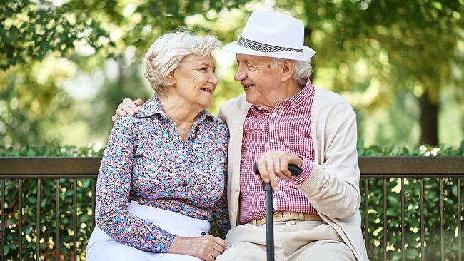 Zwei alte Personen sitzen im Grünen auf einer Bank und lächeln sich an. Der Mann hält einen Stock in einer Hand, die andere Hand hat er um die Schulter der Frau gelegt.